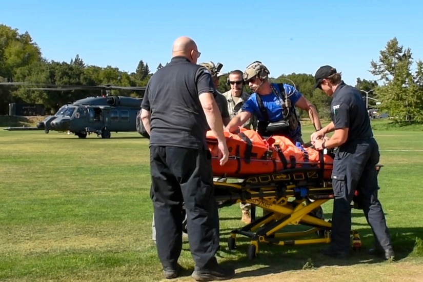 Five people secure a person to a medical transport stretcher in a grassy field with a helicopter behind them.