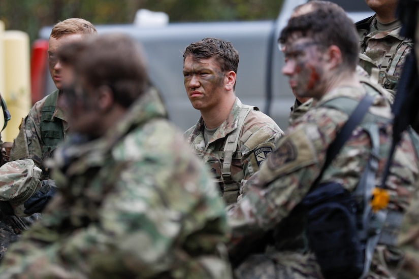 A group of soldiers wearing camouflage military uniforms and face paint, sits and listens to a person not pictured.