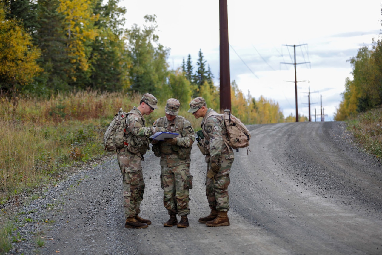 Three soldiers wearing camouflage military uniforms and backpacks stand on the side of a gravel road looking at a map attached to a clipboard.