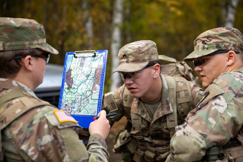 Two soldiers wearing camouflage military uniforms look at the back of a clipboard, while another soldier in similar attire holds it up while looking at a map attached to the clipboard.