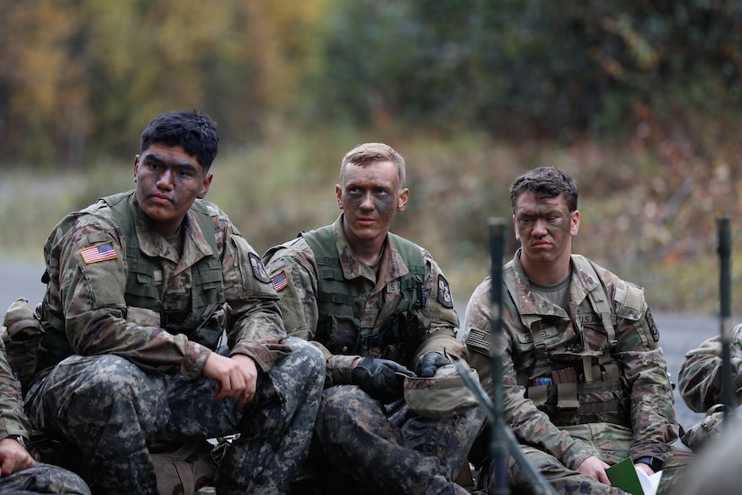 Three soldiers wearing camouflage military uniforms and face paint sit while looking to their left.