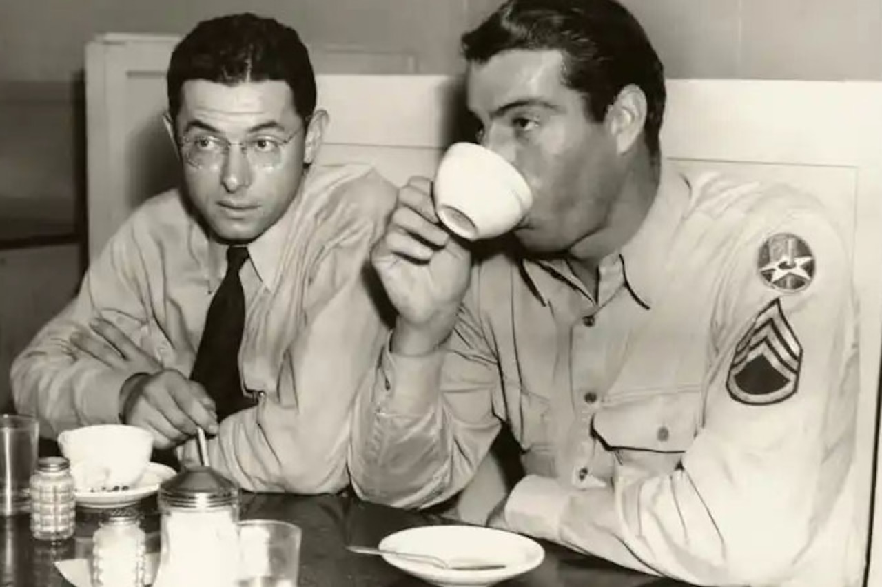 Two men wearing military uniforms sit at a table. The man on the right is drinking a cup of coffee and there is another cup on the table in front of the other man.