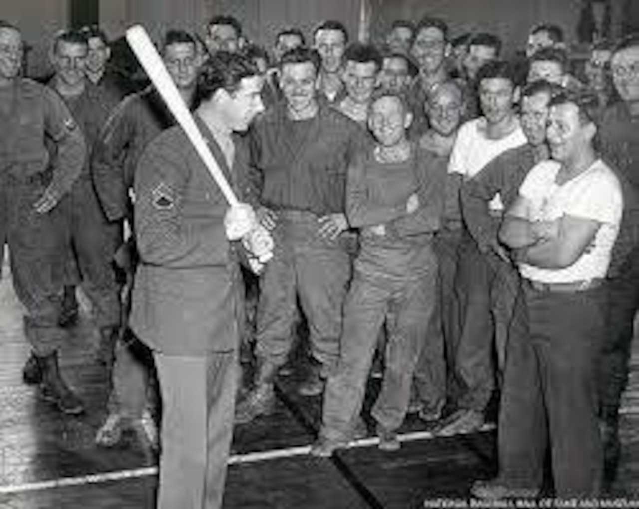 A soldier wearing a military uniform holds a baseball bat while other soldiers look on and smile.