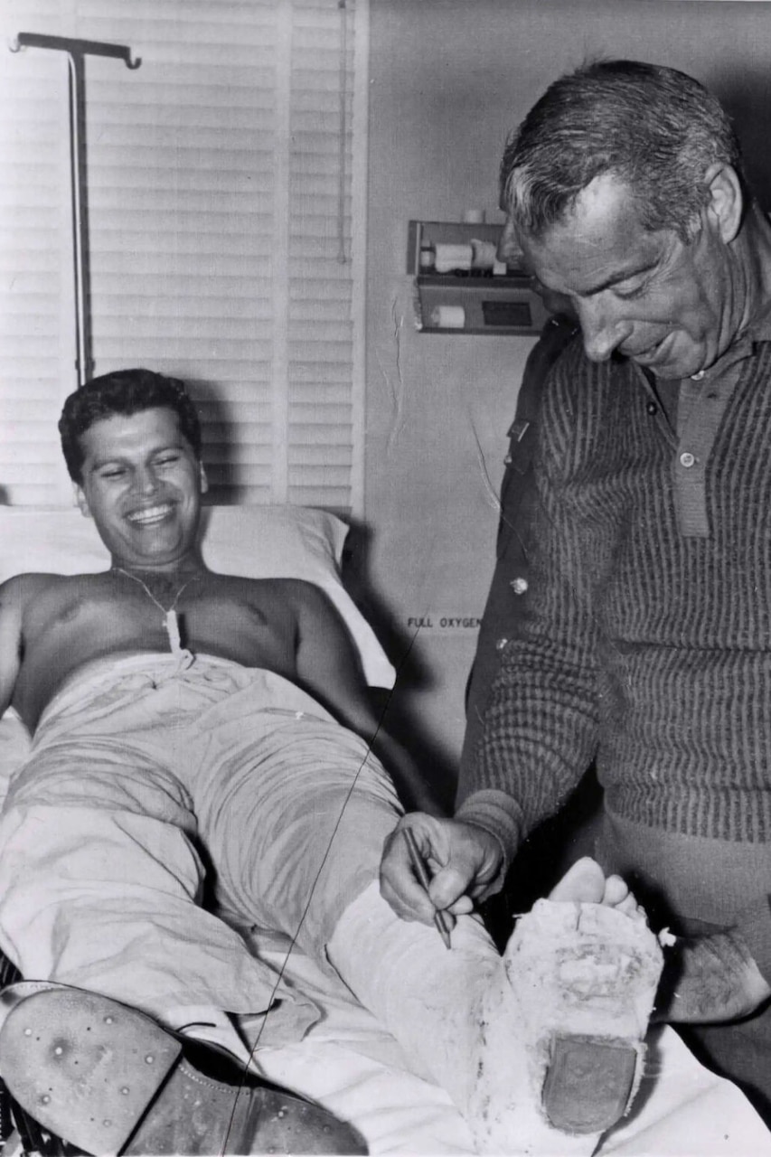A man in casual attire signs a leg cast of a man lying down on a hospital bed.