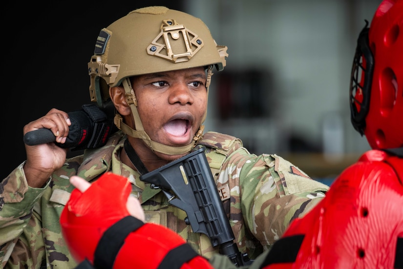 A man wearing an advanced combat helmet and camouflage military uniform swings a baton at a person wearing a red padded suit during baton combatives training.