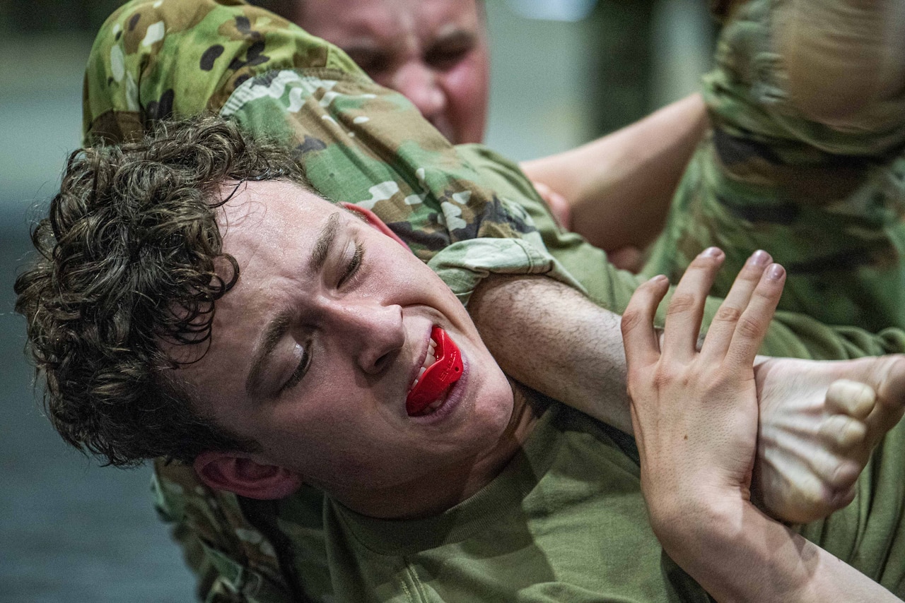 Two men wearing camouflage military uniforms wrestle during combatives training. One man has a red bite guard in his mouth.