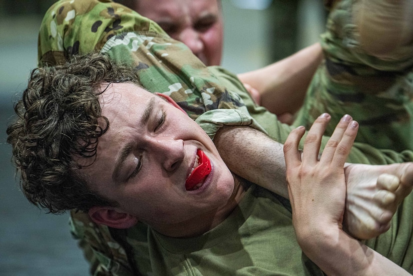 Two men wearing camouflage military uniforms wrestle during combatives training. One man has a red bite guard in his mouth.