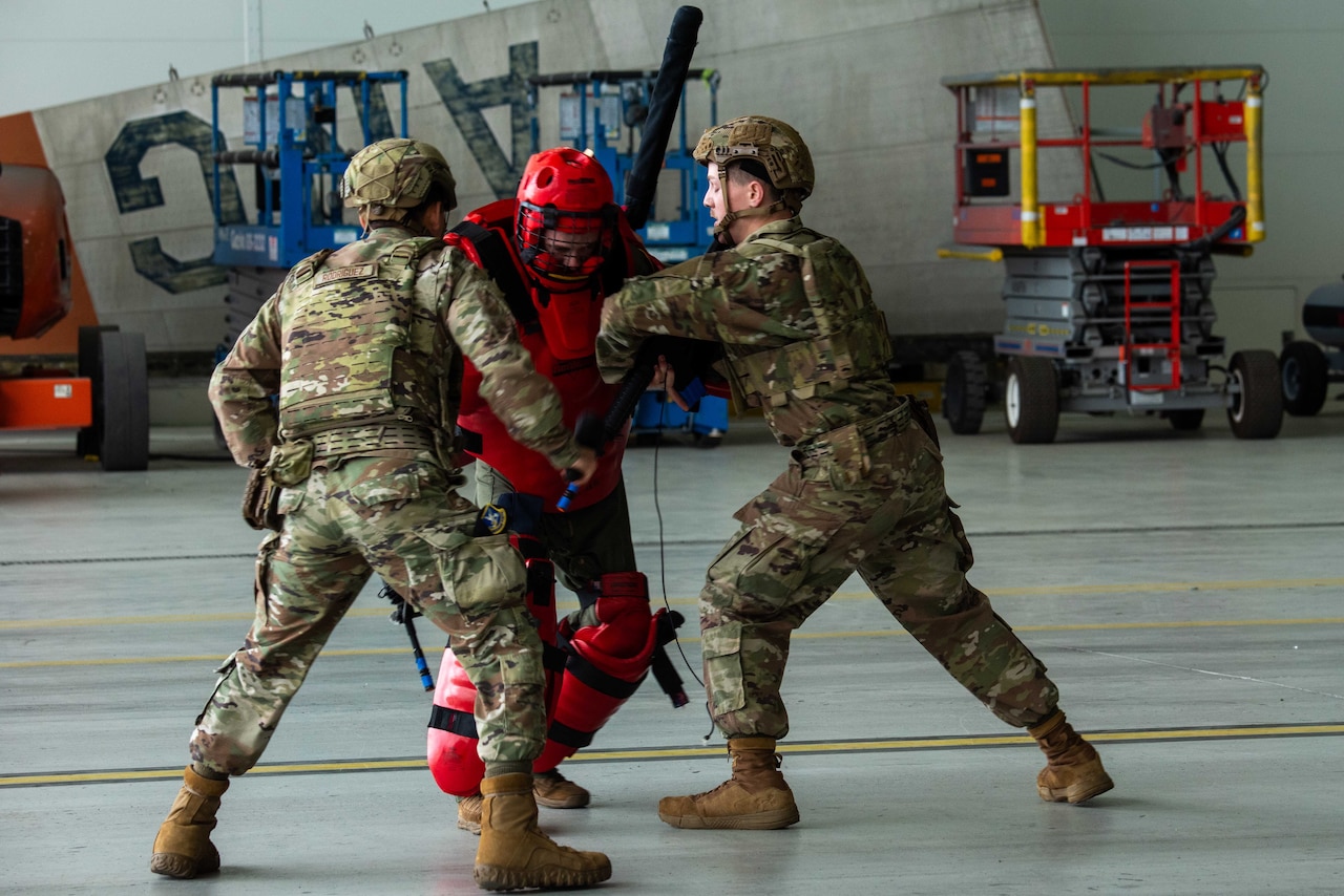 Two men wearing advanced combat helmets and camouflage military uniforms push a person dressed in a red padded suit and helmet during combatives training.