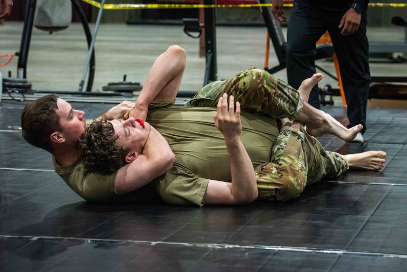 Two men dressed in camouflage military uniforms wrestle during combatives training. One man has a red bite guard in his mouth.