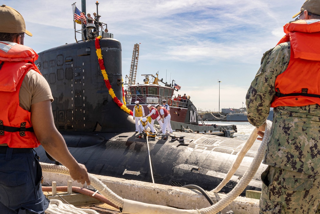 Sailors wearing life vests hold a line while standing on a port as sailors work on a submarine displaying an American flag.