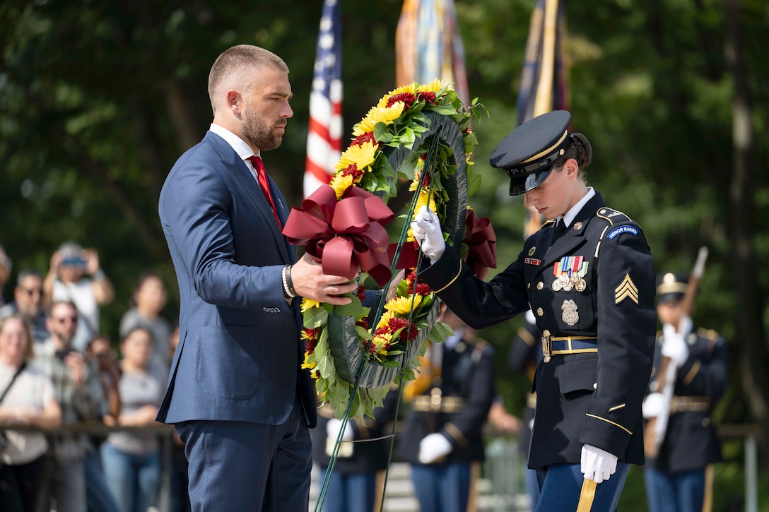 A service member looks down while holding a stand as a person in a suit holds a wreath during the day in front of a blurred crowd.