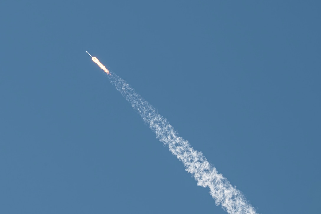 A rocket launches across a blue sky leaving behind a white streak.