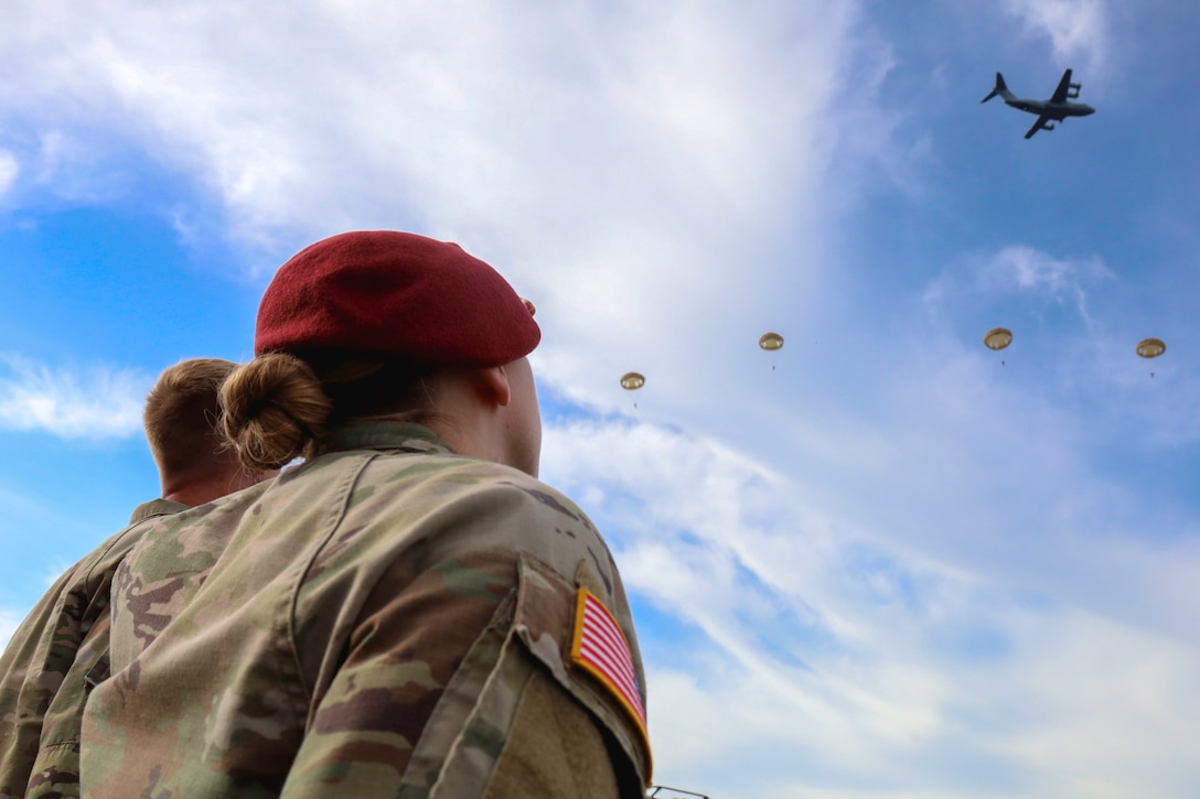 Two soldiers watch paratroopers floating down from an aircraft against a partly cloudy blue sky as seen from below.