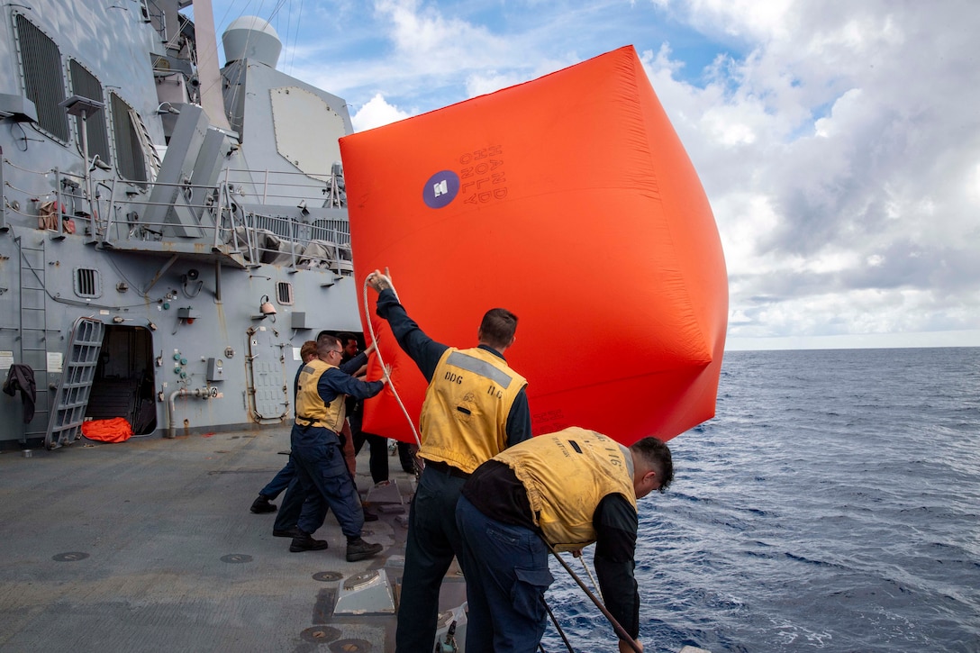 Sailors in yellow vests stand on the edge of a ship and hold up a large, orange, inflatable buoy over the ocean during the day.