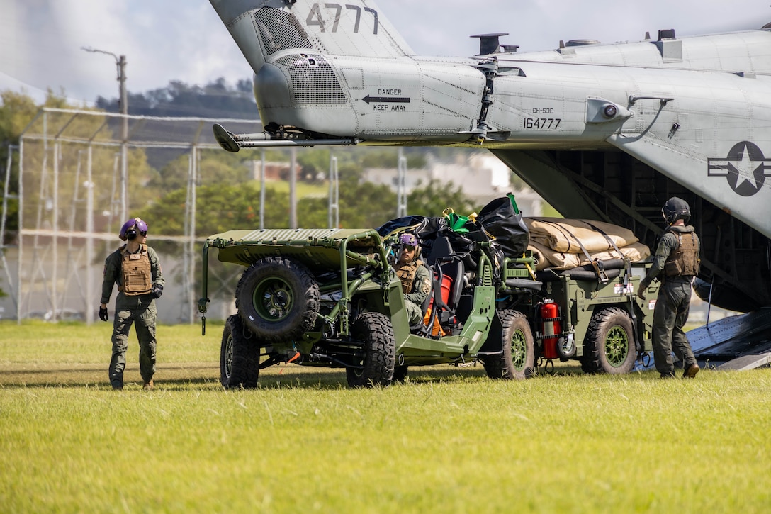 A Marine drives a loaded ATV out of an aircraft as two Marines stand nearby in a field during the day.
