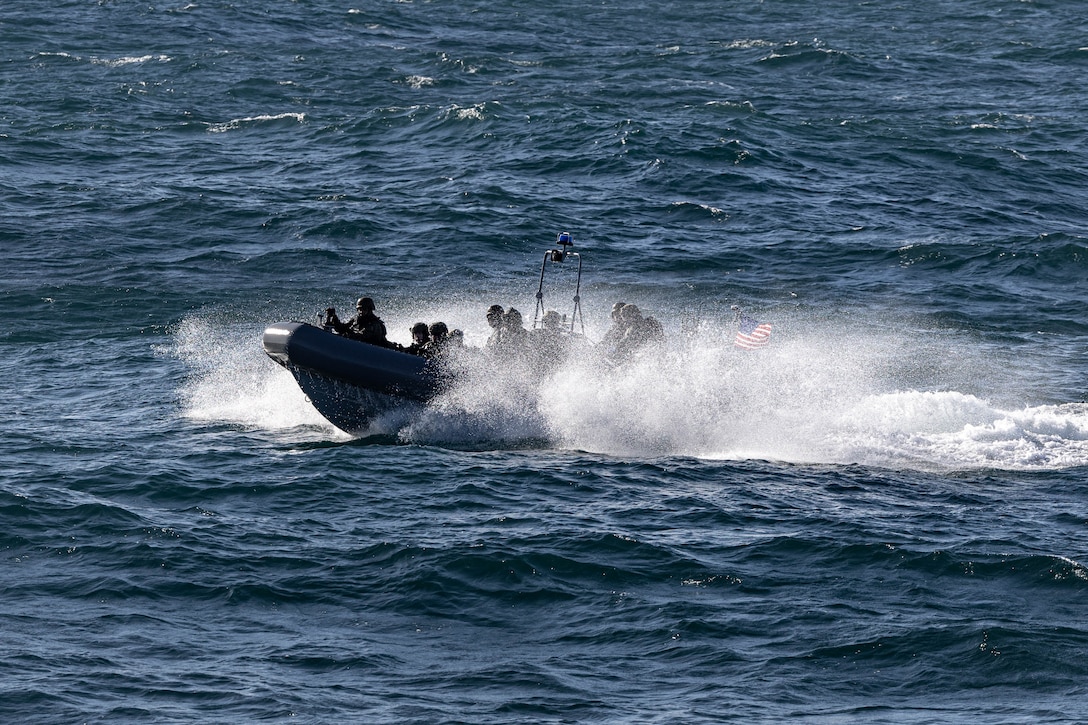 Sailors in a small boat with an American flag on the back motor through choppy ocean waves during the day.