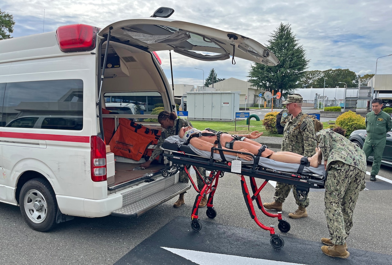 Three sailors wearing camouflage military uniforms load a medical mannequin into the back of an emergency vehicle.