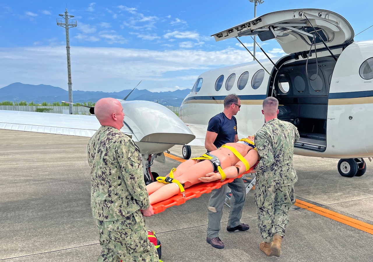 Two sailors wearing camouflage military uniforms and a man in civilian attire load a medical mannequin onto a medical jet.