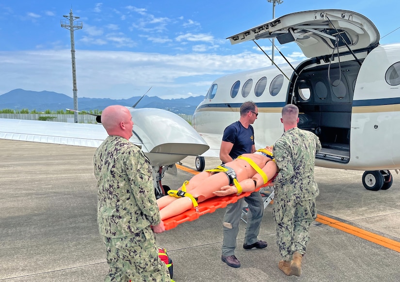 Two sailors wearing camouflage military uniforms and a man in civilian attire load a medical mannequin onto a medical jet.
