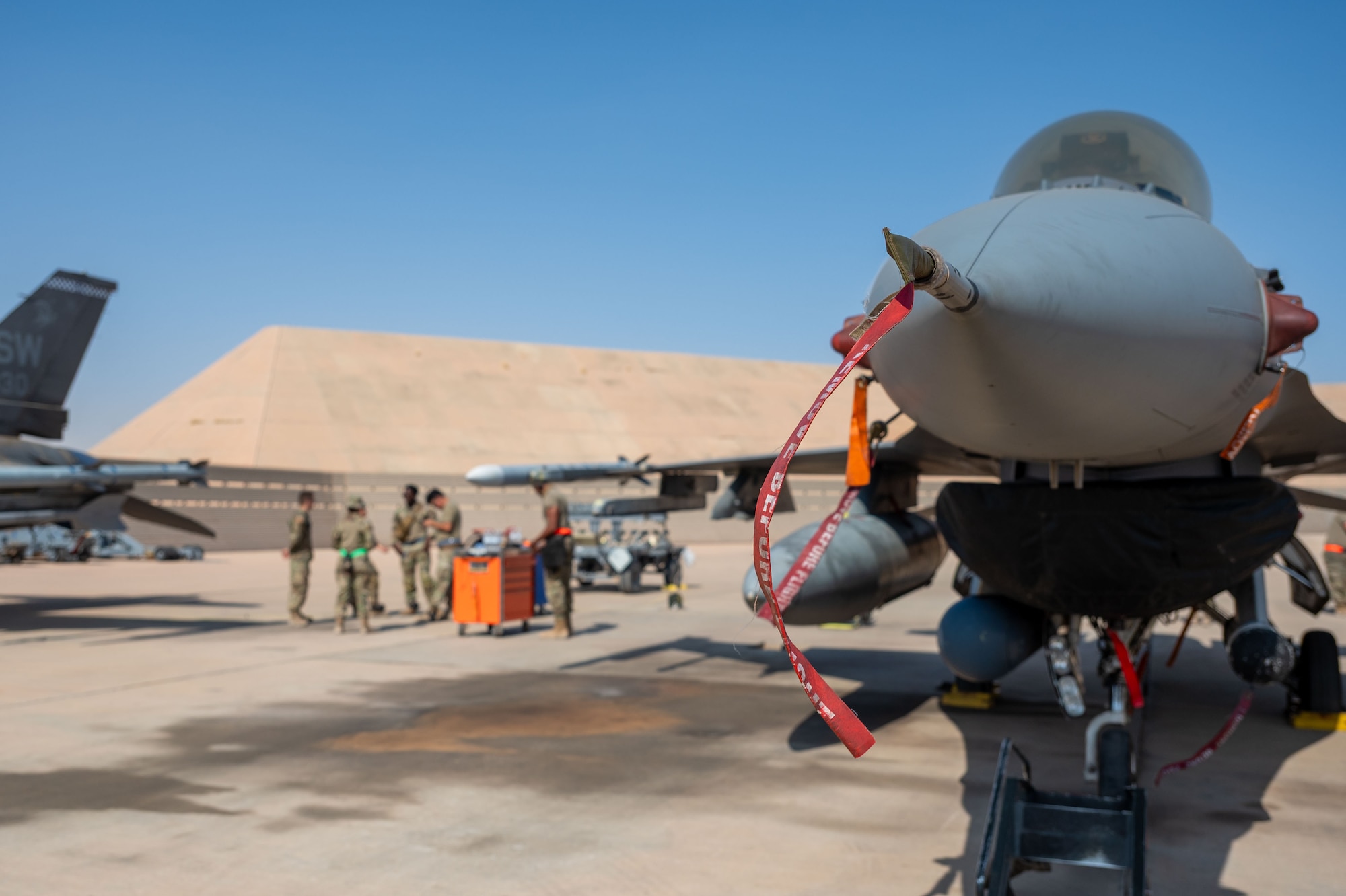 The nose of an F-16 Fighting Falcon is in focus in front of Airmen debriefing.