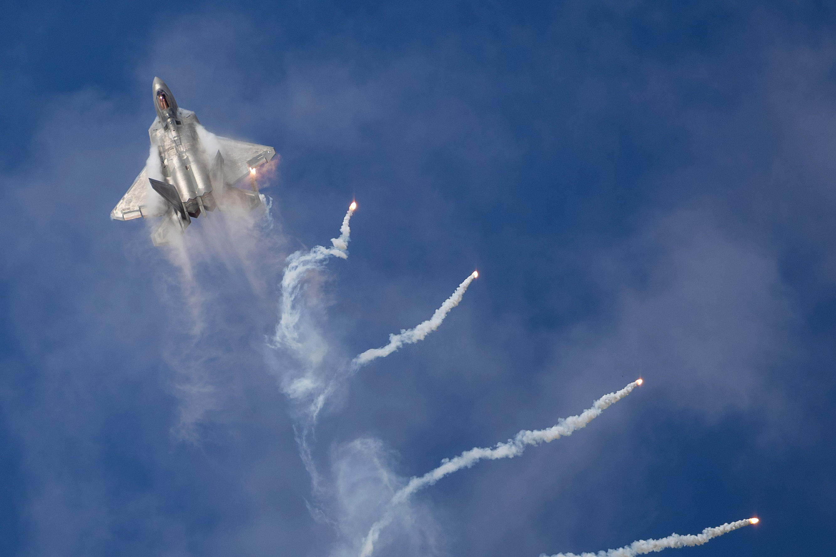 An aircraft flies above four flares and trails of smoke against a blue sky.