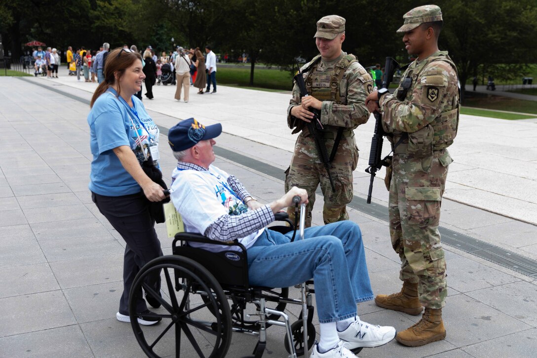 Two guardsmen in camouflage military uniforms and holding rifles talk to two people in civilian clothing, one sitting in a wheelchair and holding a cane, on a paved walkway, with other people, trees and a grassy area in the background during daytime.