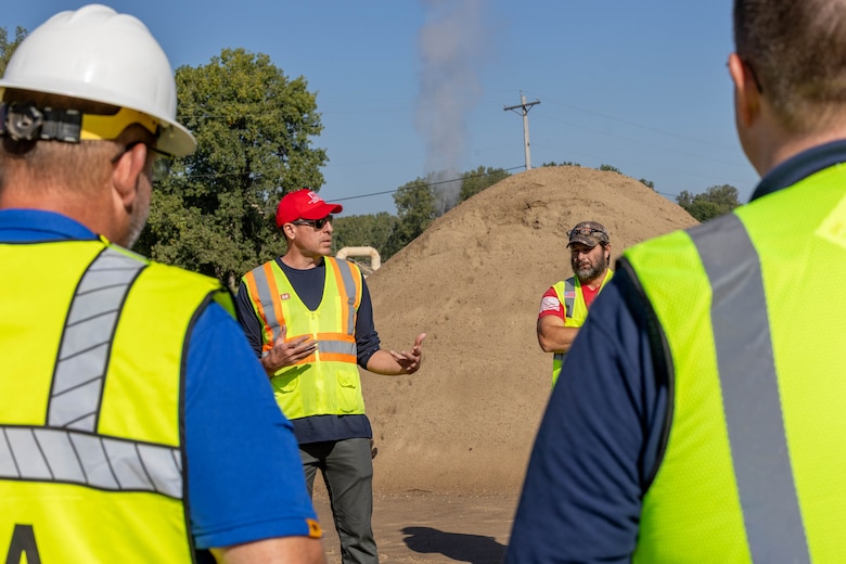A man in a red had and yellow safety vest speaks to a crowd of people in yellow safety vests with a mound of sand in the background.