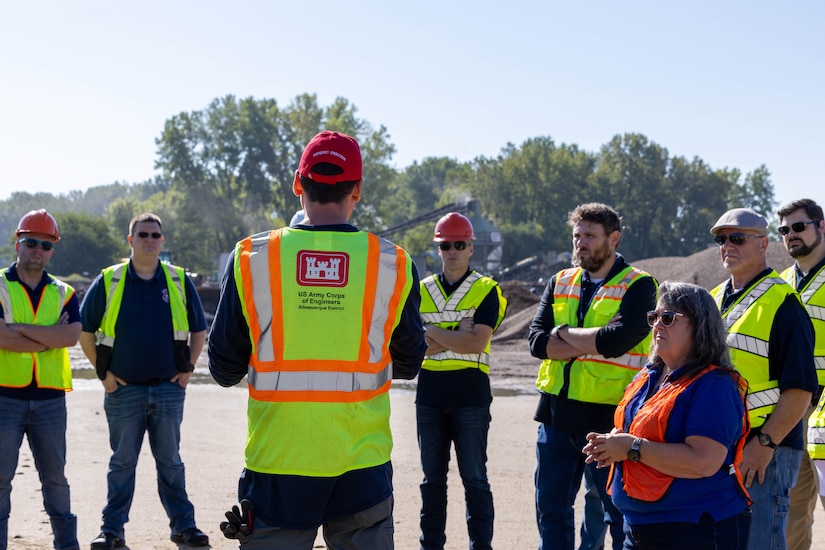 A man in a red had and yellow safety vest speaks in front of a group of people in yellow safety vests.