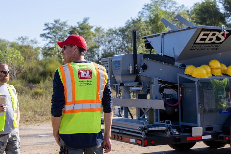 A man in a yellow safety vest and red hat stands in front of a grey sandbag machine.