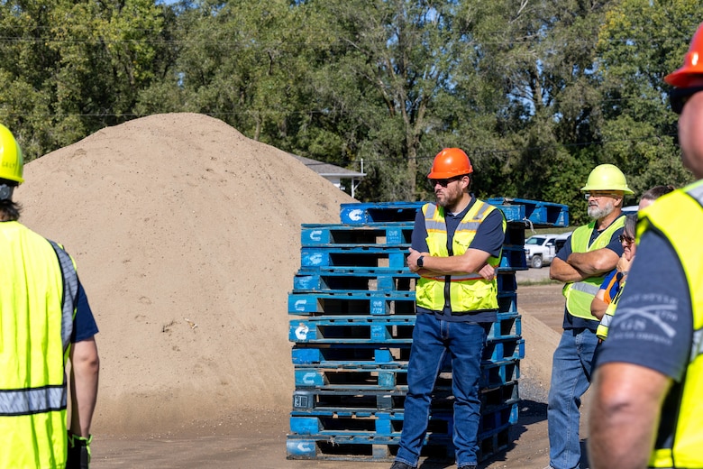 Four men in yellow safety vests and yellow and orange hard hats stand in front of blue wood pallets and a mound of sand.