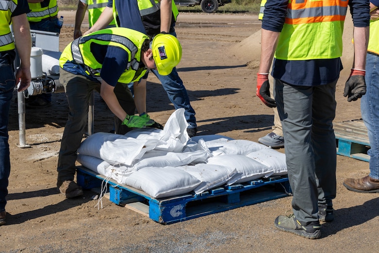 A man in a yellow safety vest and hardhat places a white bag full of sand on top of other white bags of sand on top of a blue pallet with sand in the background.