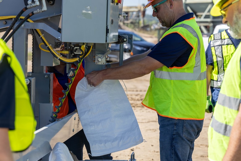 A man in a yellow safety vest holds a white bag under a grey sandbag machine.