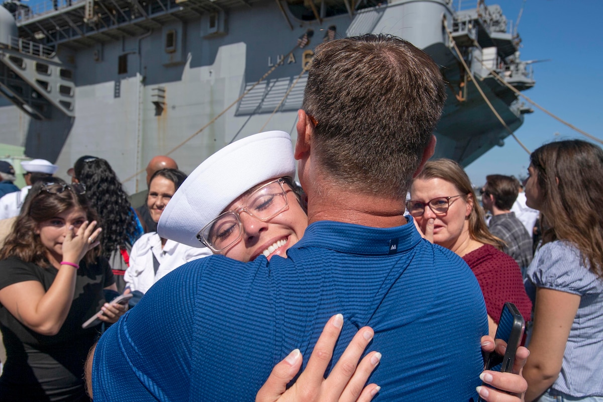 A sailor smiles while hugging a loved one as emotional family members watch with a ship in the background.