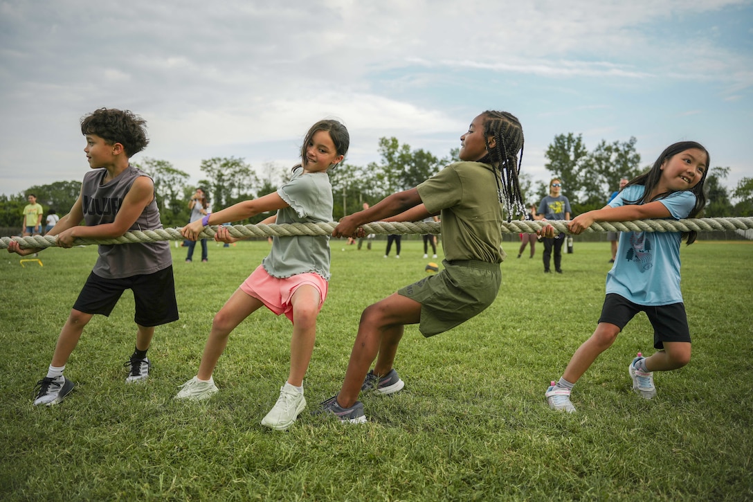 Four children pull a rope while playing tug-of-war on a field during the day.