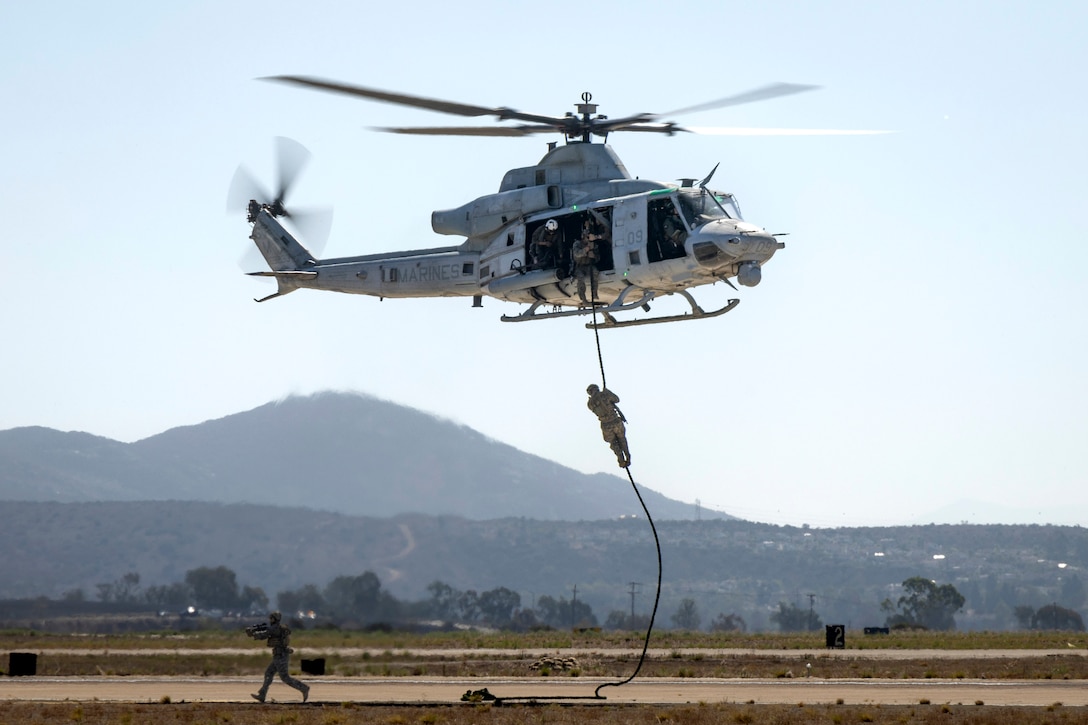 A Marine rappels from a rope attached to a hovering helicopter as a fellow Marine watches from the open doorway during the day with mountains in the background.