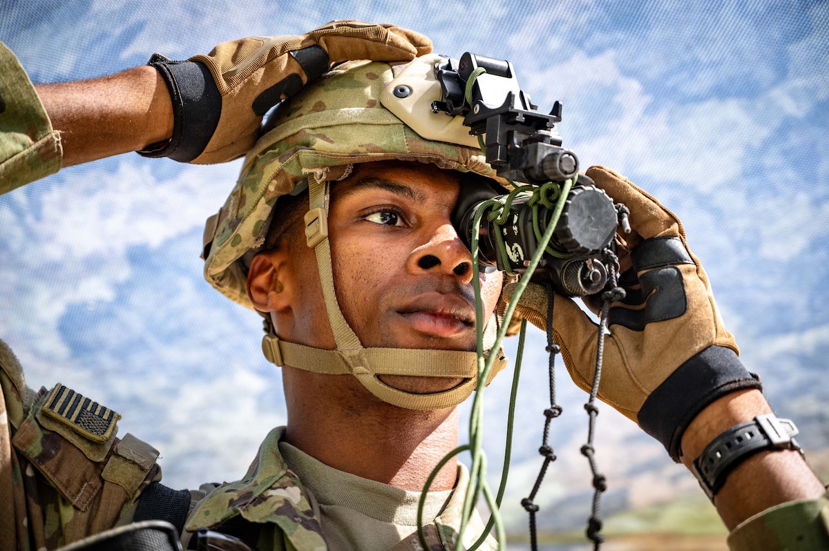 A soldier places night-vision goggles on their helmet under a blue sky, as seen from close-up.
