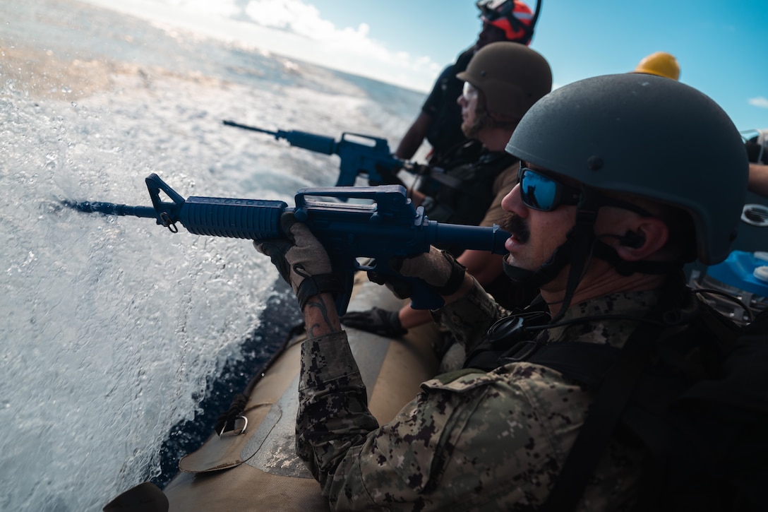 Sailors in helmets hold service weapons as they sit in an inflatable boat on the ocean.
