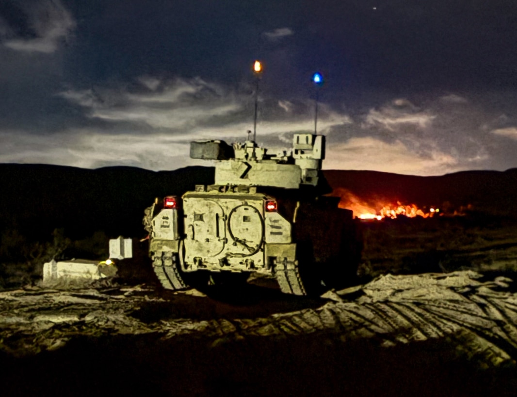 A tank is parked on a dirt path under a night sky as fires burn in the background.
