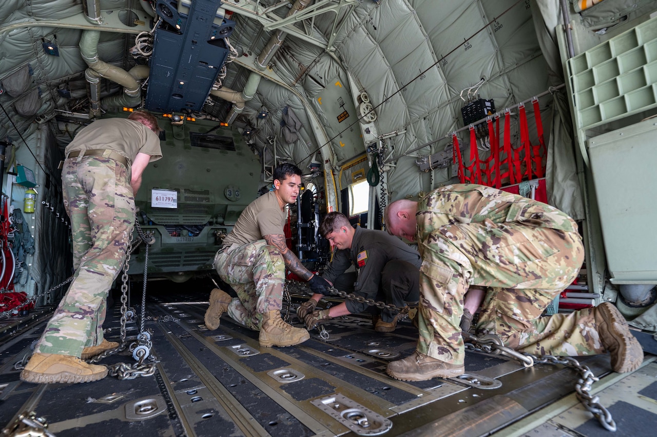 Three men wearing camouflage military uniforms and another man wearing coveralls attach chains to the floor of a military cargo aircraft. The chains are securing a military vehicle inside the aircraft that is in the background.
