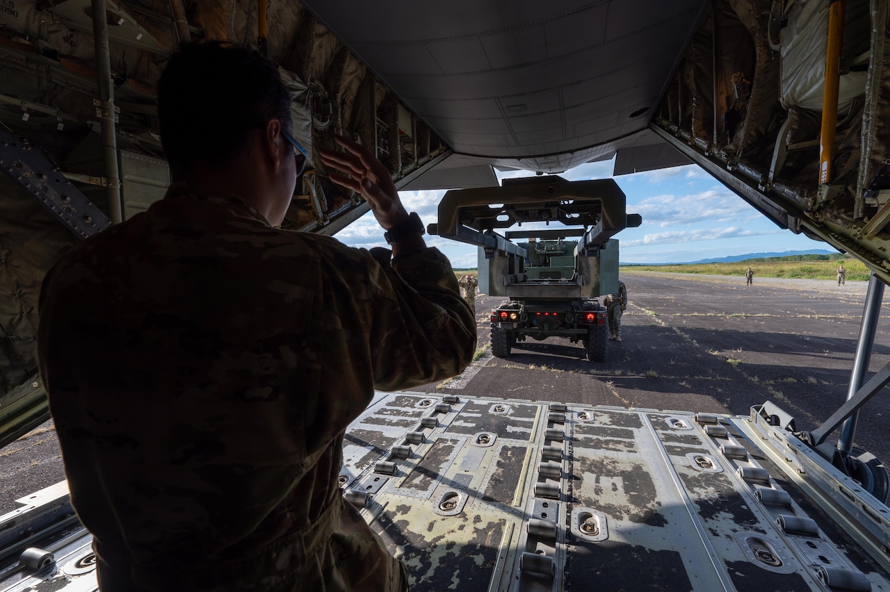 A man wearing a camouflage military uniform guides a military vehicle into the back a large military cargo aircraft. There are four other service members in similar attire standing outside the aircraft on the tarmac.