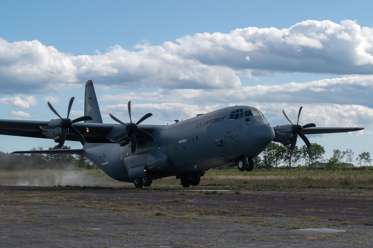 A large gray military cargo aircraft takes off from a runway. There is a field and trees in the background.