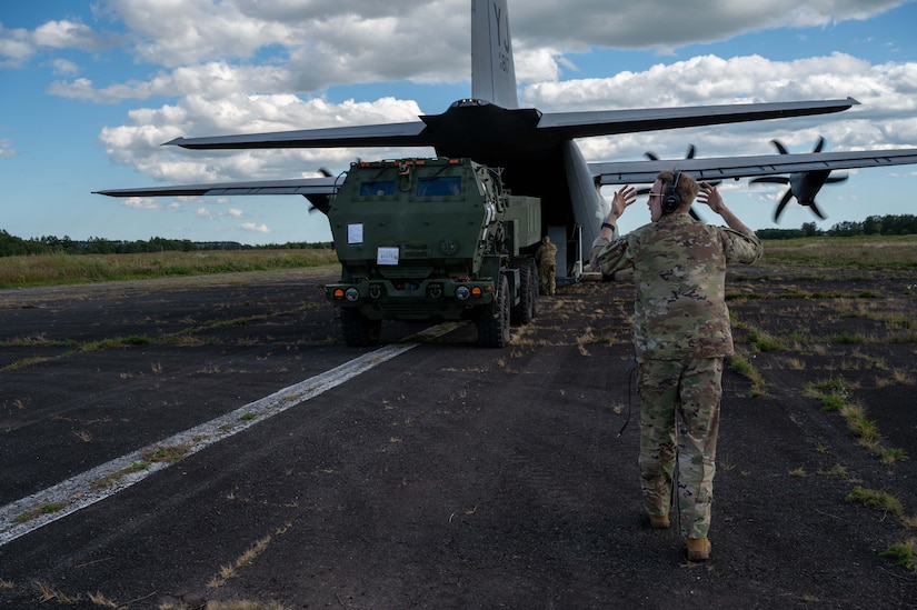 A man wearing a camouflage military uniform walks backwards on a tarmac as he guides a large green military vehicle out of a military cargo aircraft.