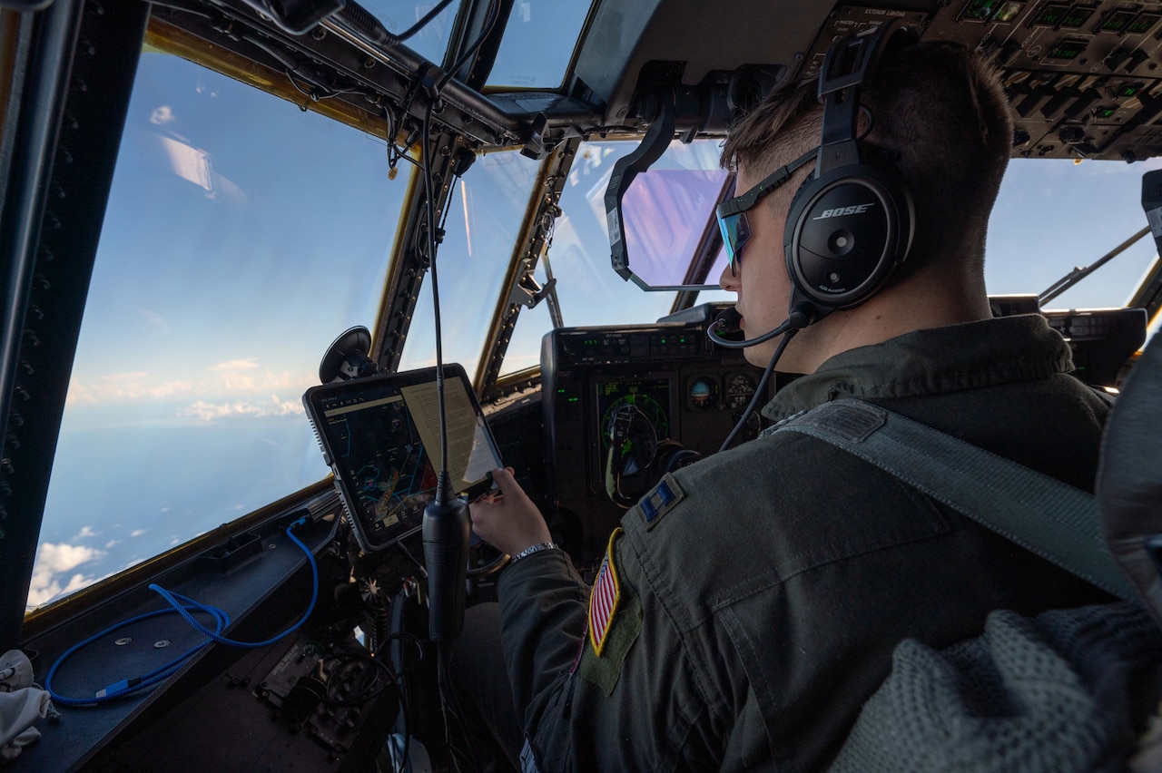 A man wearing a flight suit, sunglasses and a headset looks at a tablet screen while sitting in the cockpit of a military cargo aircraft. The aircraft is flying, and you can see clouds outside the windows of the cockpit.