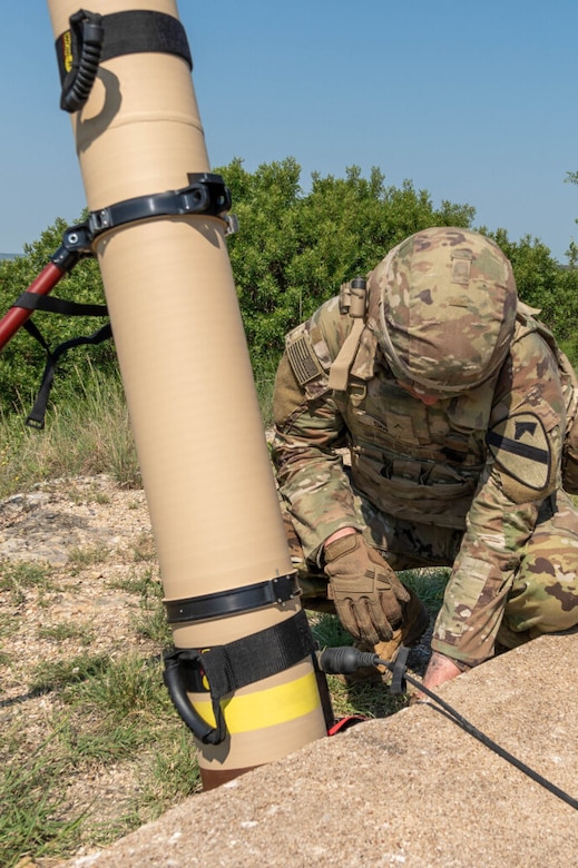 A person wearing a camouflage military uniform sets up a weapons system outside in a desert environment.