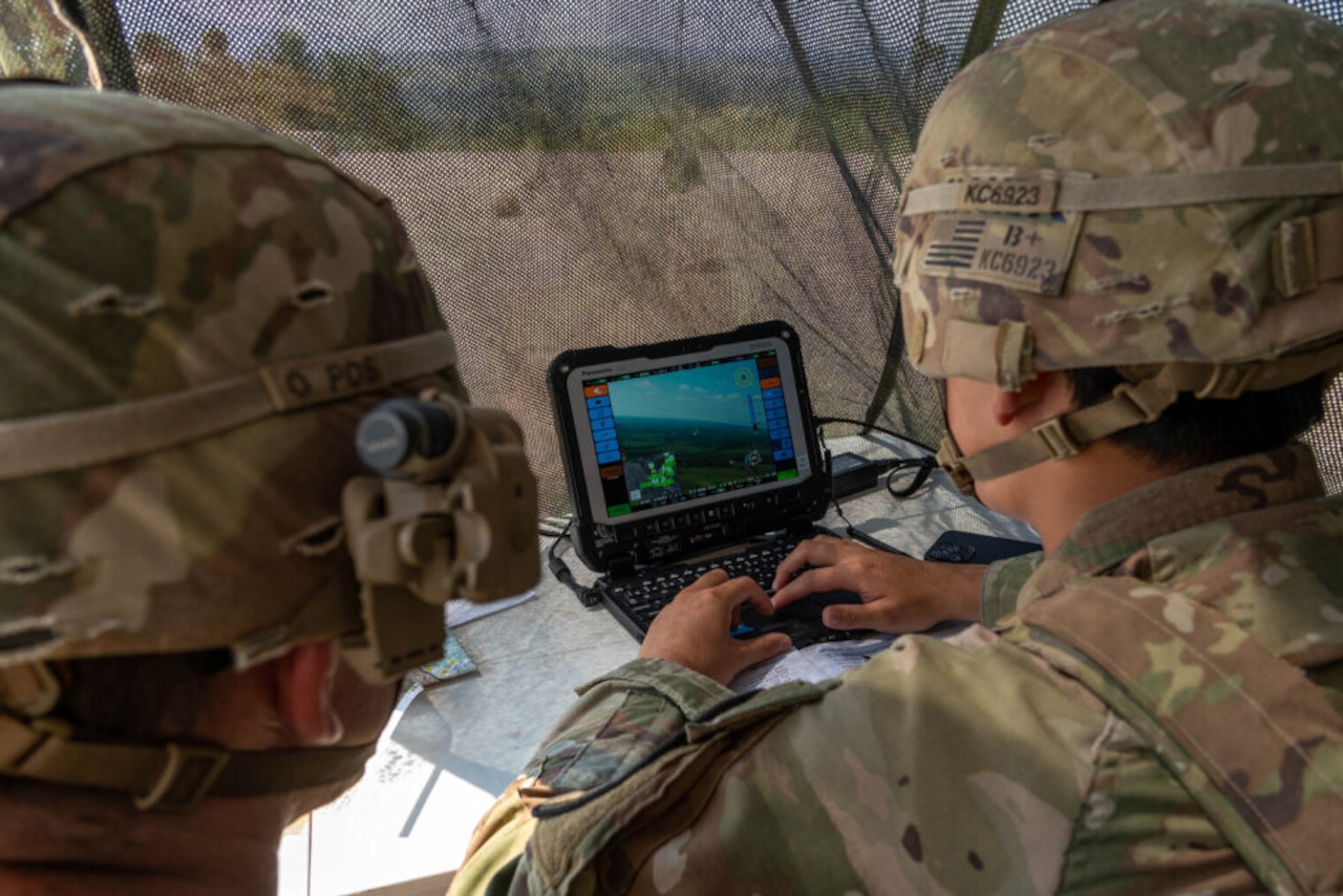 Two people wearing camouflage military uniforms and helmets look at a small screen under a camouflage-netted tent.