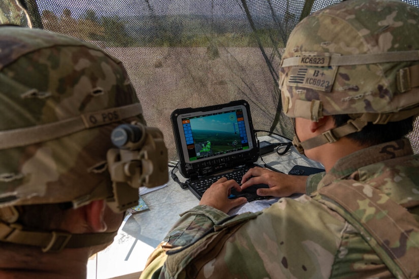 Two people wearing camouflage military uniforms and helmets look at a small screen under a camouflage-netted tent.