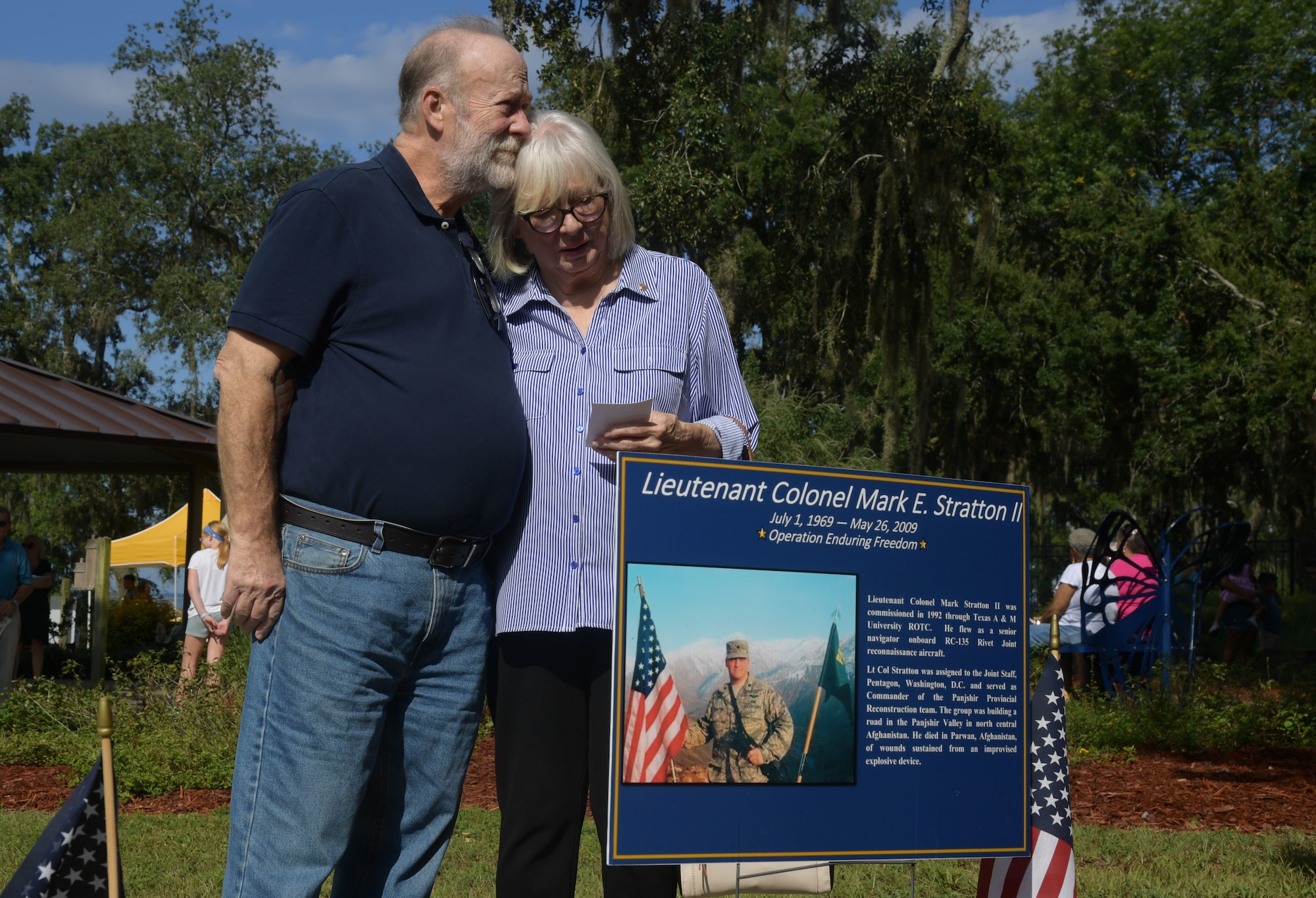 A couple stands behind a memorial poster while holding each other.