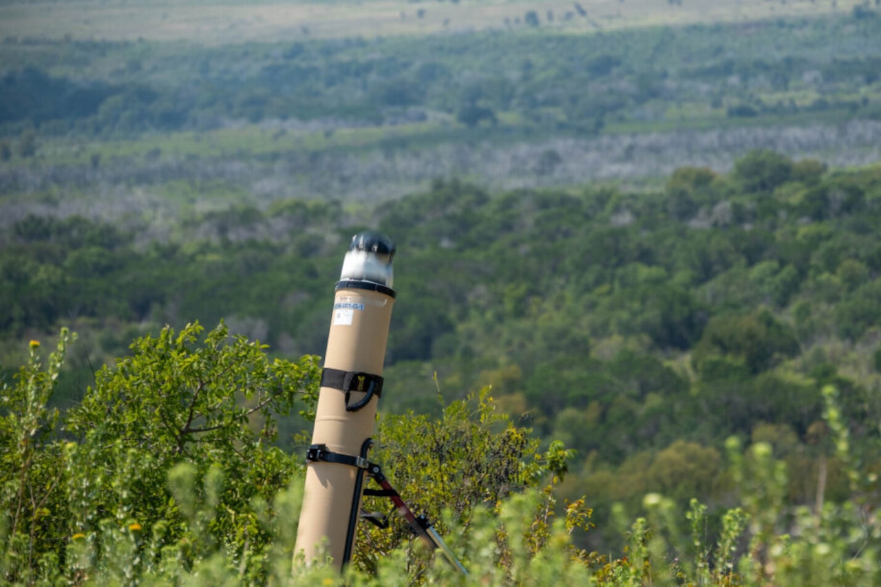 A tube-like military weapons system sits outside in a semi-wooded area.