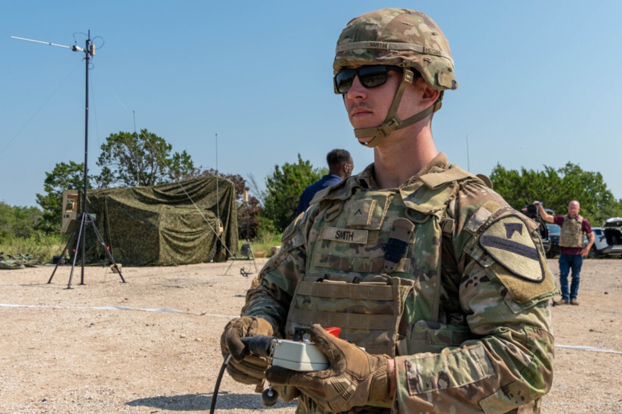 A person wearing sunglasses, a helmet and a camouflage military uniform holds an electronic switch outside in a desert environment. In the background are a camouflage tent and other personnel.