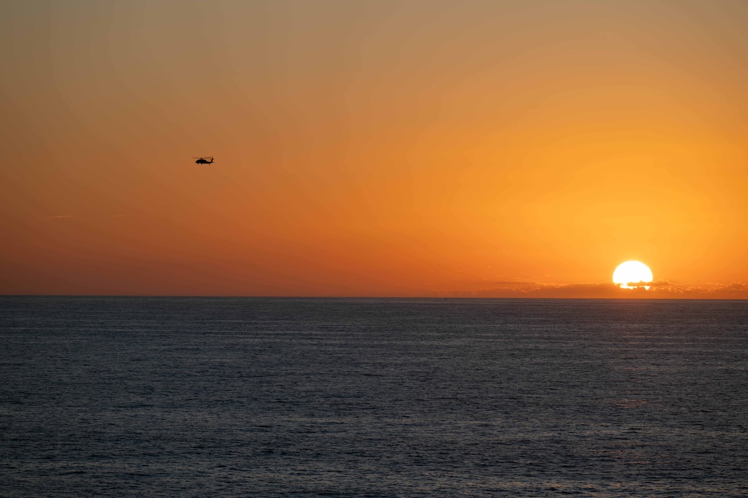 A helicopter flies above the ocean against an orange sky while the sun nears the horizon to the right.
